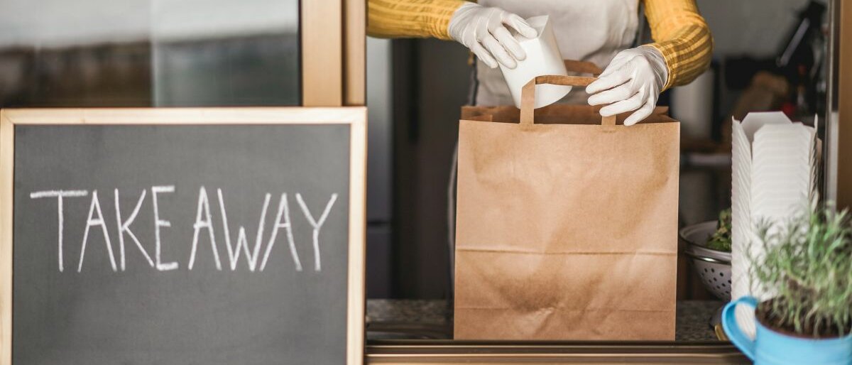Eine Frau steht hinter einer Durchreiche und packt eine Papiertüte mit Take-Away-Essen.