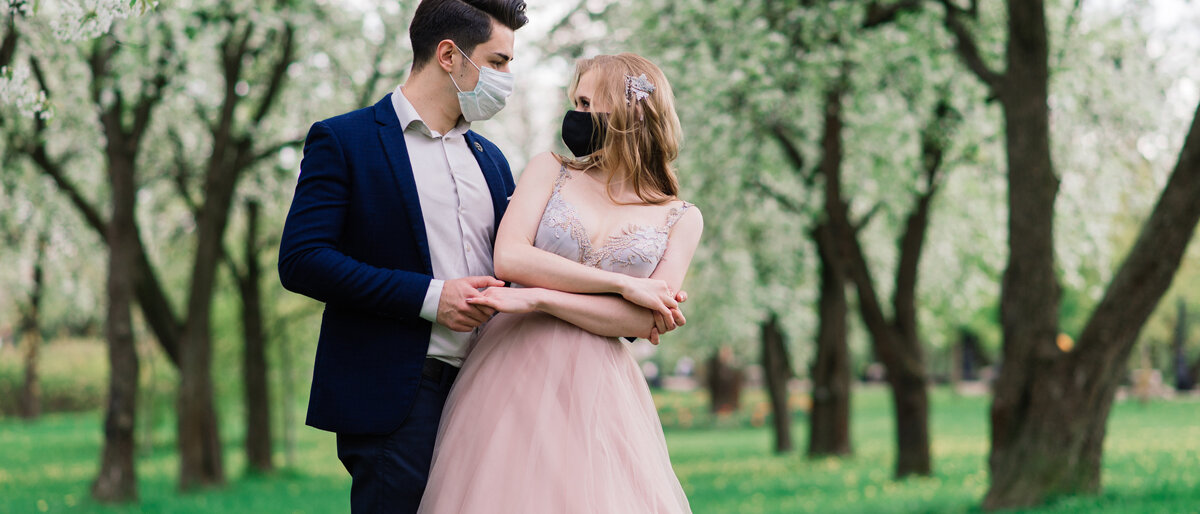 Ein junges Paar in Abendkleidung posiert in einer Kirschblütenallee unt trägt Alltagsmasken.