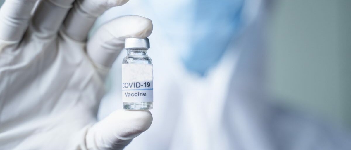 Jemand mit Schutzkleidung und Einweghandschuhen hält einen COVID-19-Impfstoff in der Hand.