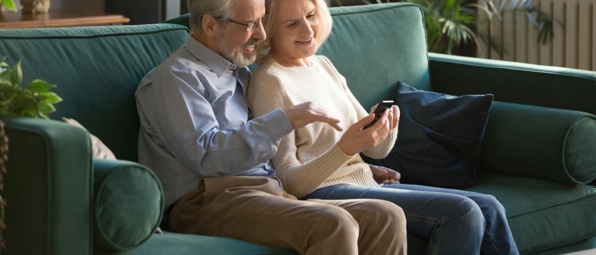 Ein älteres Paar sitzt auf der Couch mit einem Smartphone und reden miteinander..