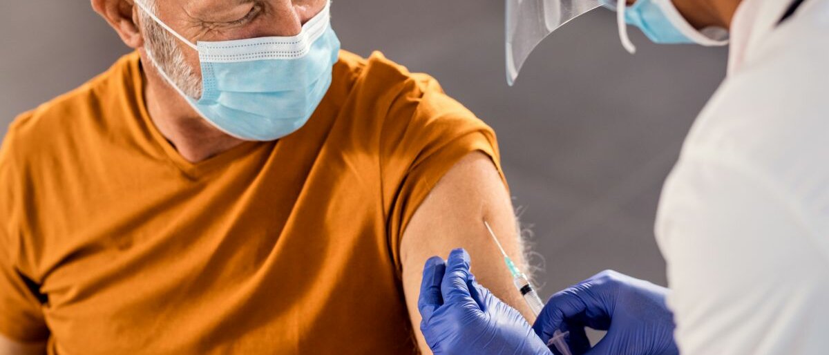 Eine Frau impft einen älteren Herren. Beide tragen eine Mund-Nasen-Bedeckung.