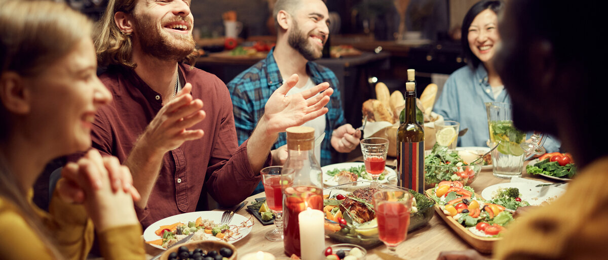 Eine Gruppe junger Menschen sitzt gemeinsam am Tisch, isst Gemüseaufläufe und lacht.