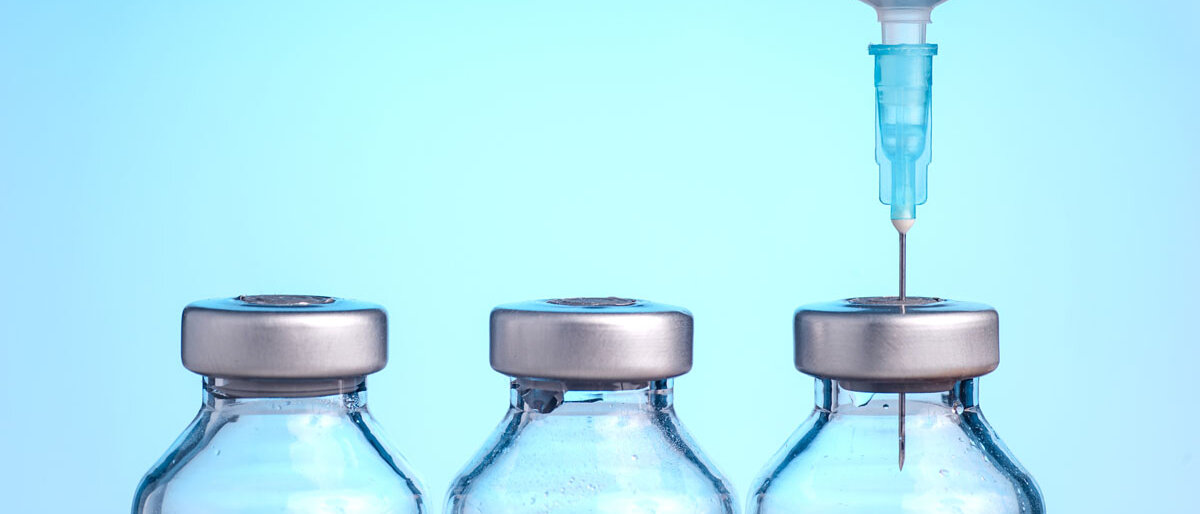 Drei Impfstoffampullen stehen nebeneinander, aus der rechten wird mit einer Spritze der Inhalt aufgezogen.