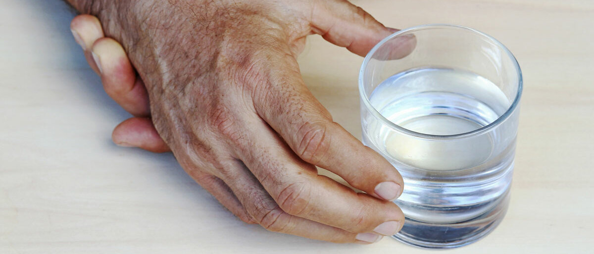 Hand greift nach Wasserglas, wird von anderer Hand gestützt, weil sie zittert