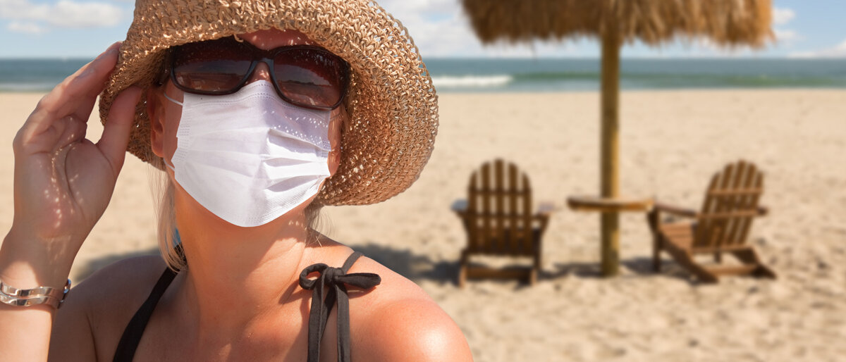 Eine Frau am Strand trägt Sonnenhut, Sonnenbrille und Mund-Nasen-Maske.