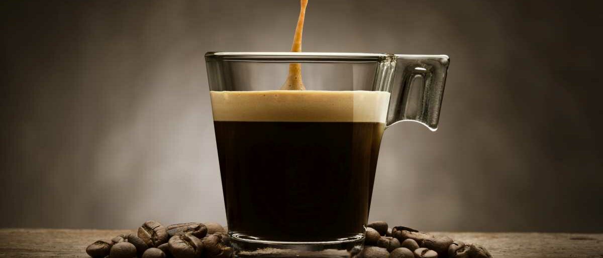 Um eine Glastasse mit Espresso darin liegen einige Kaffeebohnen.