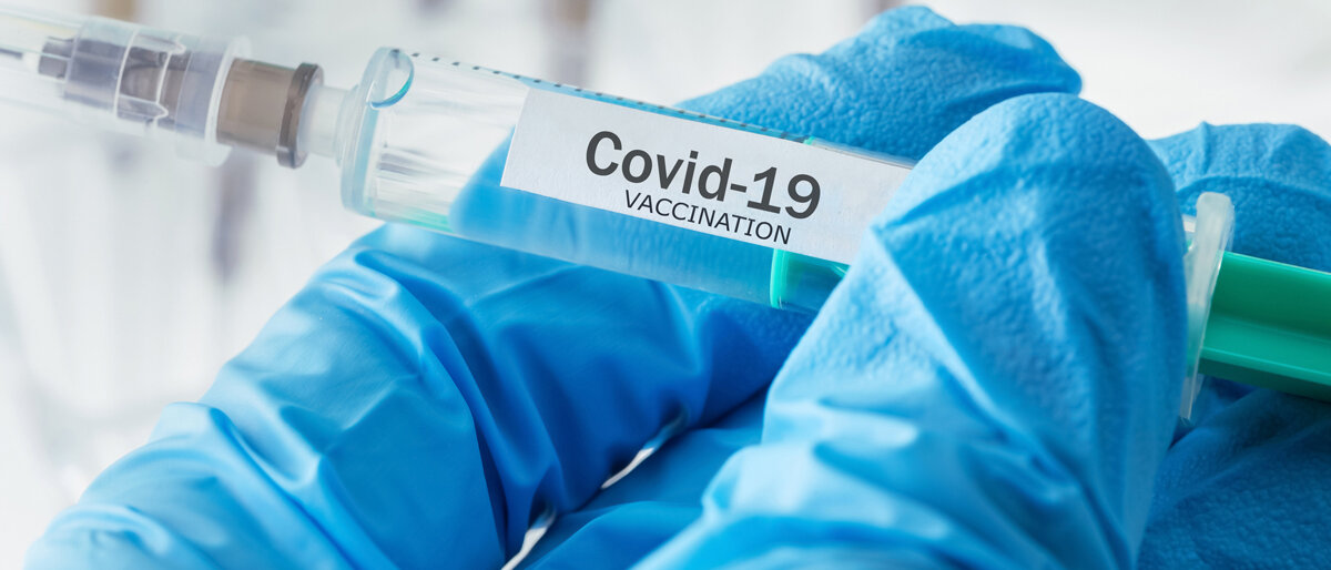 Eine Hand, die blaue Handschuhe trägt, hält eine Spritze mit der Aufschrift "Covid-19 Vaccination".