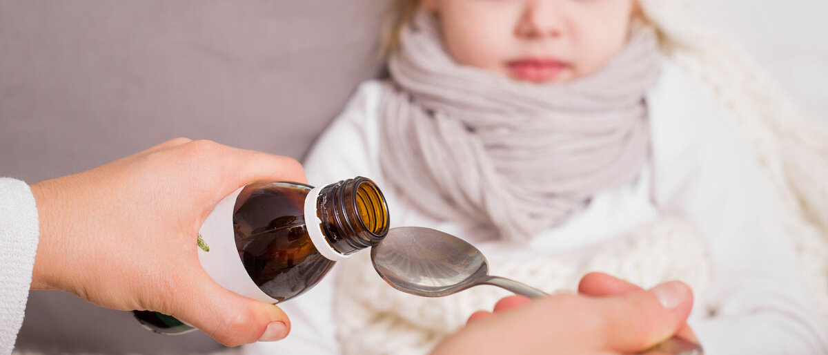 Krankes Kind im Bett, bekommt gerade einen Arznei-Saft von seiner Mutter