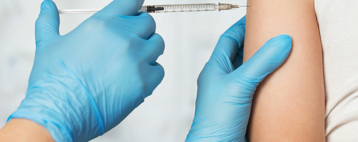 Impfung Eine behandschuhte Hand injiziert einer Person in weißem Top eine Spritze in den Oberarm.