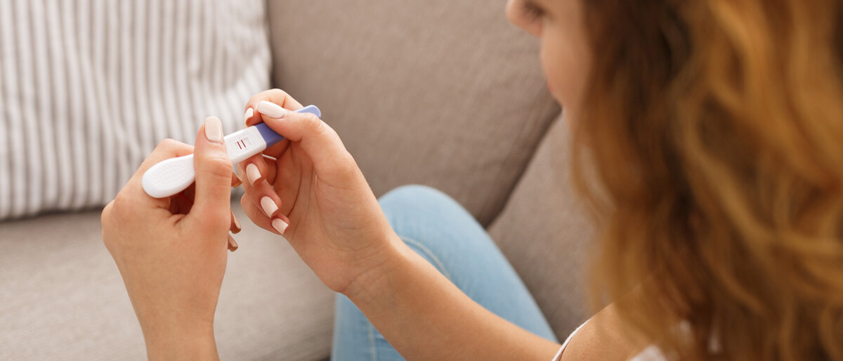 Eine junge Frau sitzt auf dem Sofa und betrachtet einen positiven Schwangerschaftstest.