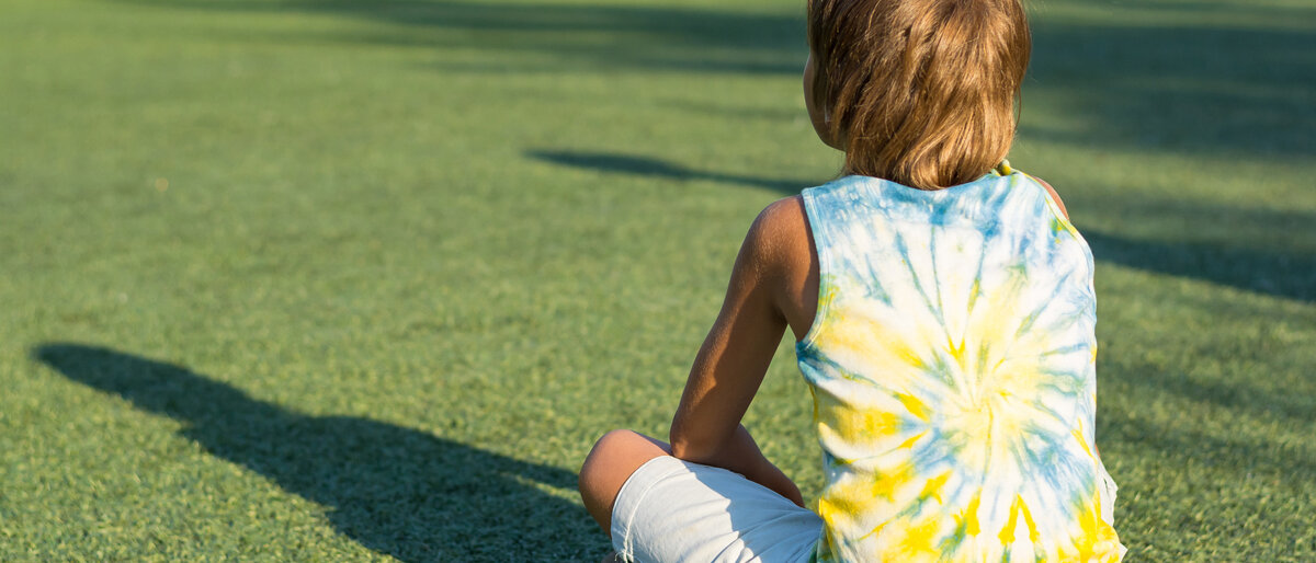 Ein Junge sitzt auf einem Fußballfeld auf dem Boden und schaut anderen Kindern beim Spielen zu.