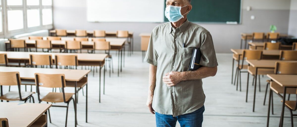 Ein leeres Klassenzimmer, indem nur ein Lehrer mit Mundschutz steht.