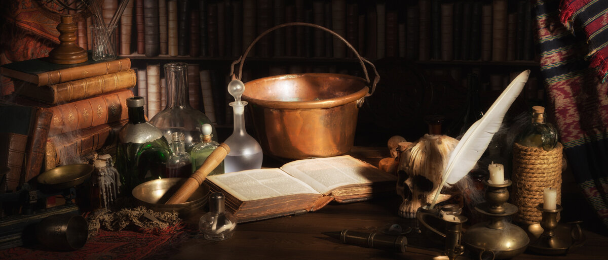 Auf einem dunklen Holzschreibtisch liegt ein aufgeschlagenes, dickes Buch. Darum herum stehen ein Kupferkessel, einige Glasphiolen, ein Totenschädel und historische Instrumente.