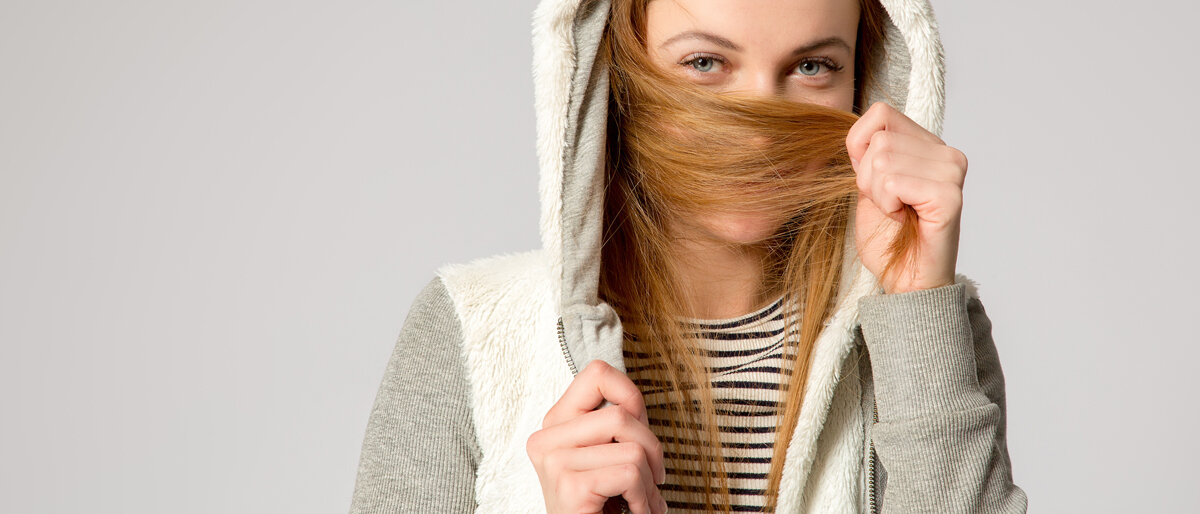 Eine junge Frau trägt einen Kapuzenpulli und hält sich ihre langen Haare vor die untere Gesichtshälfte.