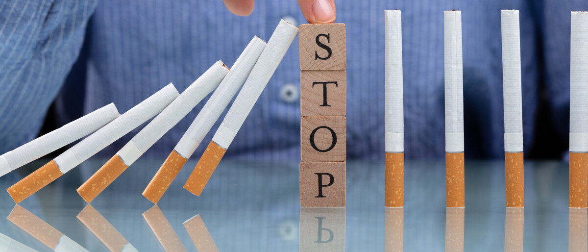 Eine Person hält einen Holzklotz mit der Aufschrift "STOP" zwischen mehrere Zigaretten, um zu verhindern, dass sie wie Dominosteine umfallen.