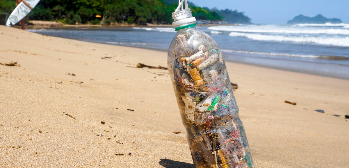 Eine mit Kippen gefüllte Plastikflasche steht an einem Strand