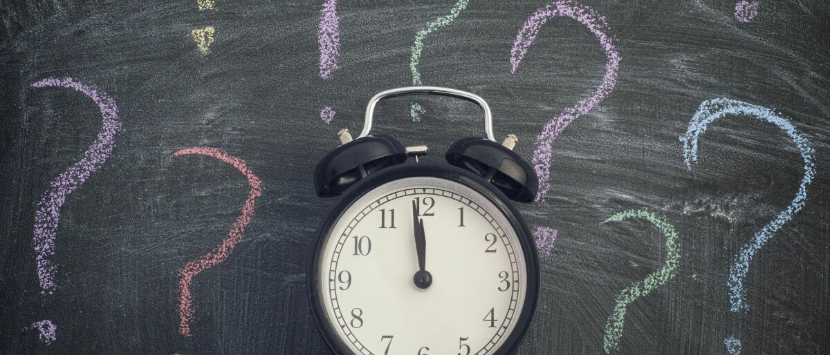 Wecker Wecker steht vor einer Tafel mit bunten Fragezeichen.