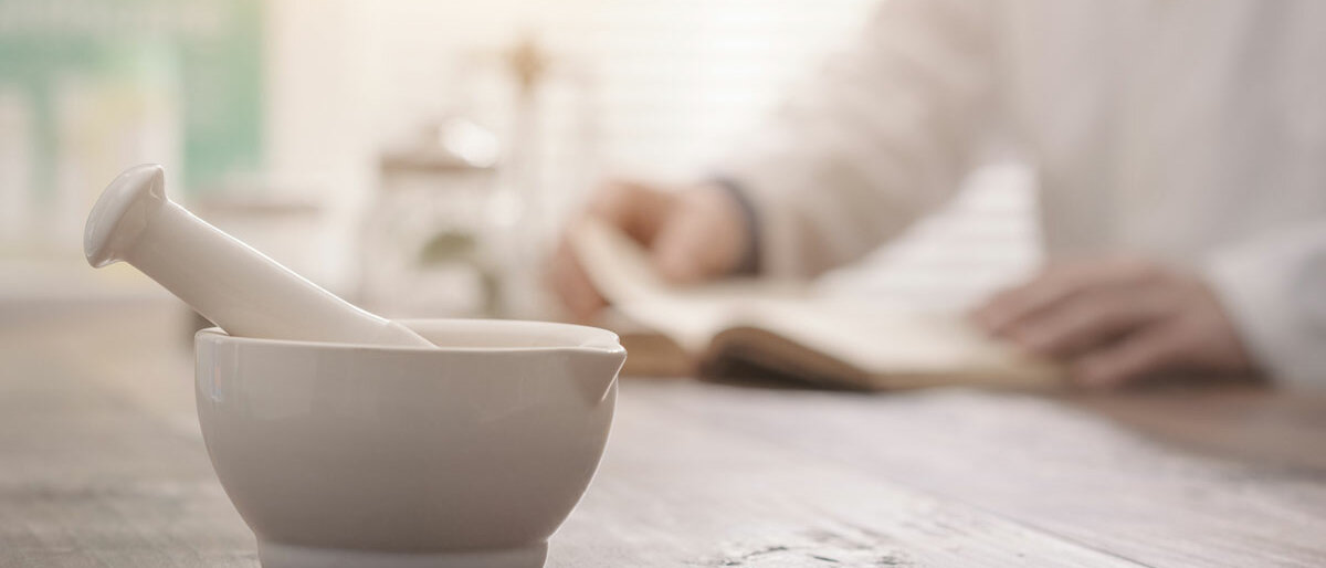 Im Vordergrund eine Rührschale mit Pistill, im Hintergrund eine Hand, die etwas aufschreibt