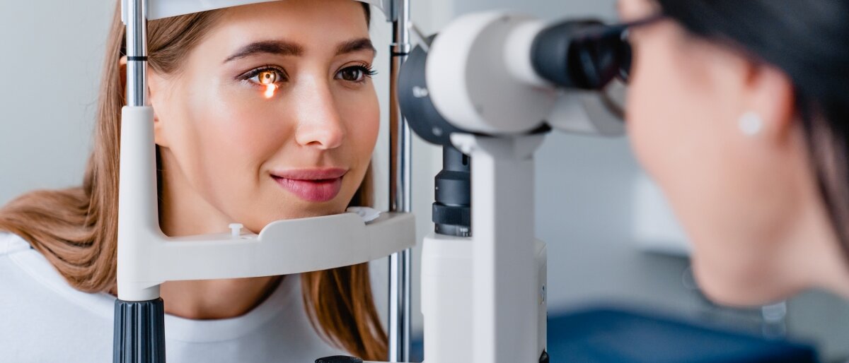 Eine junge Frau hat ihr Kinn auf eine Halterung gelegt und ihr Auge wird mit dem Spaltlampenmikroskop untersucht.
