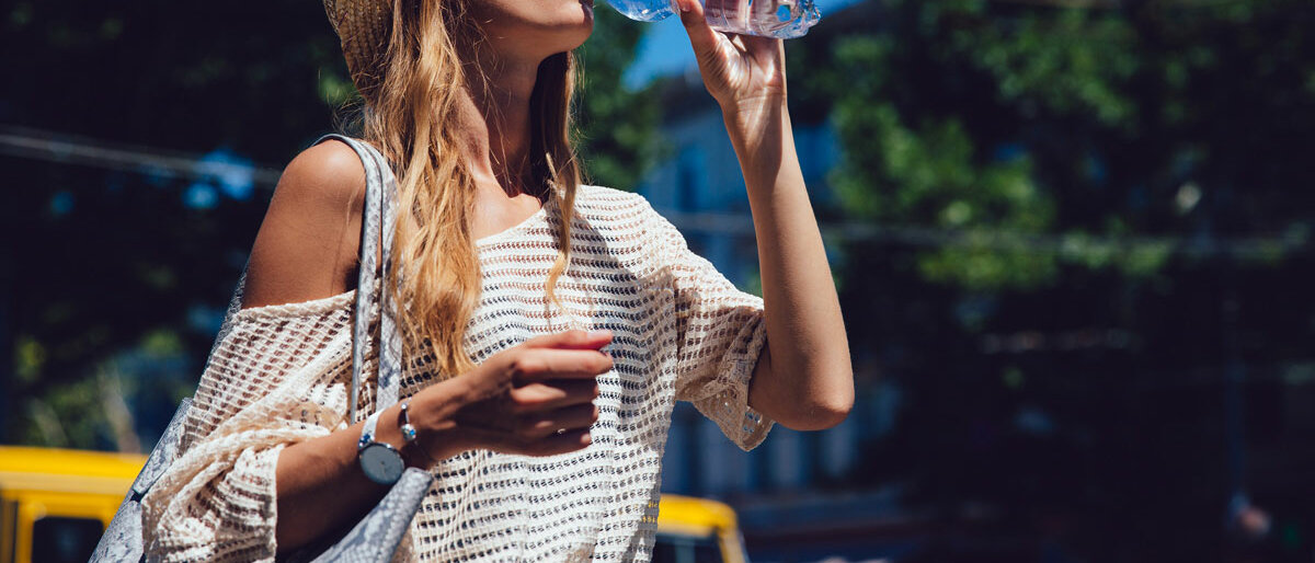 Eine junge Frau  mit Sonnenhut trinkt Wasser aus einer Flasche während die Sonne scheint