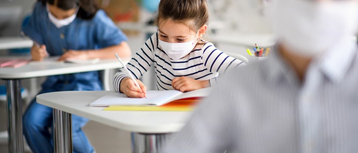 Ein Mädchen mit Mundschutz sitzt in der Schule an einem Tisch und schreibt etwas.