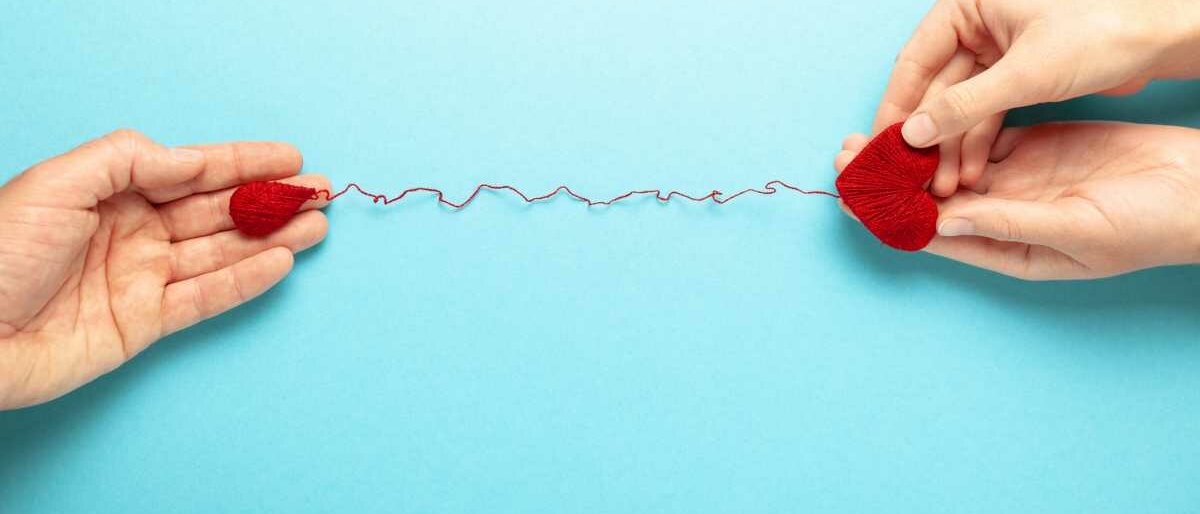 Blutspende Eine Person hält ein Herz aus Wolle in der Hand, das über einen Faden mit einem Woll-Blutstropfen in der Hand einer zweiten Person verbunden ist.