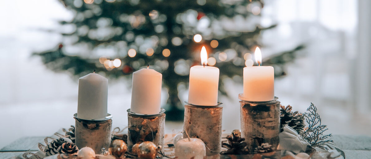 Im Hintergrund steht ein Weihnachtsbaum, im Vordergrund steht ein Adventsgesteck, auf dem zwei Kerzen brennen.