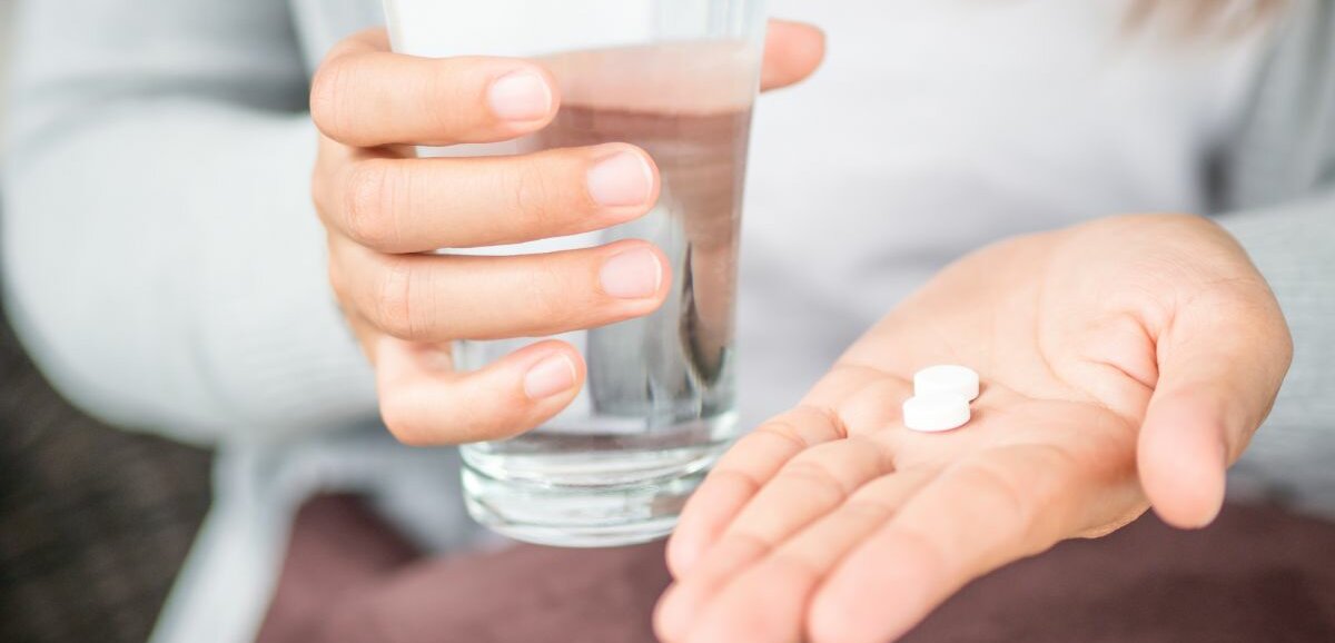 Eine Frau hält ein Glas Wasser in der einen, zwei weiße Tabletten in der anderen Hand.