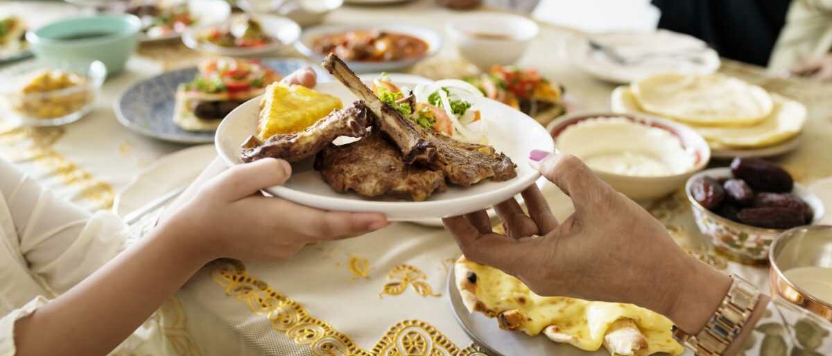 Eine Familie sitzt am reich gedeckten Tisch, eine Frau reicht einer anderen einen Teller mit Lammkeulen und Bazlama.