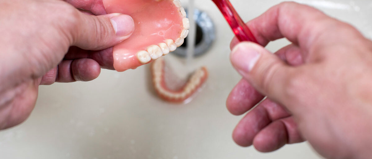 Eine Zahnprothese wird im Waschbecken unter laufendem Wasser mit einer Bürste gereinigt.