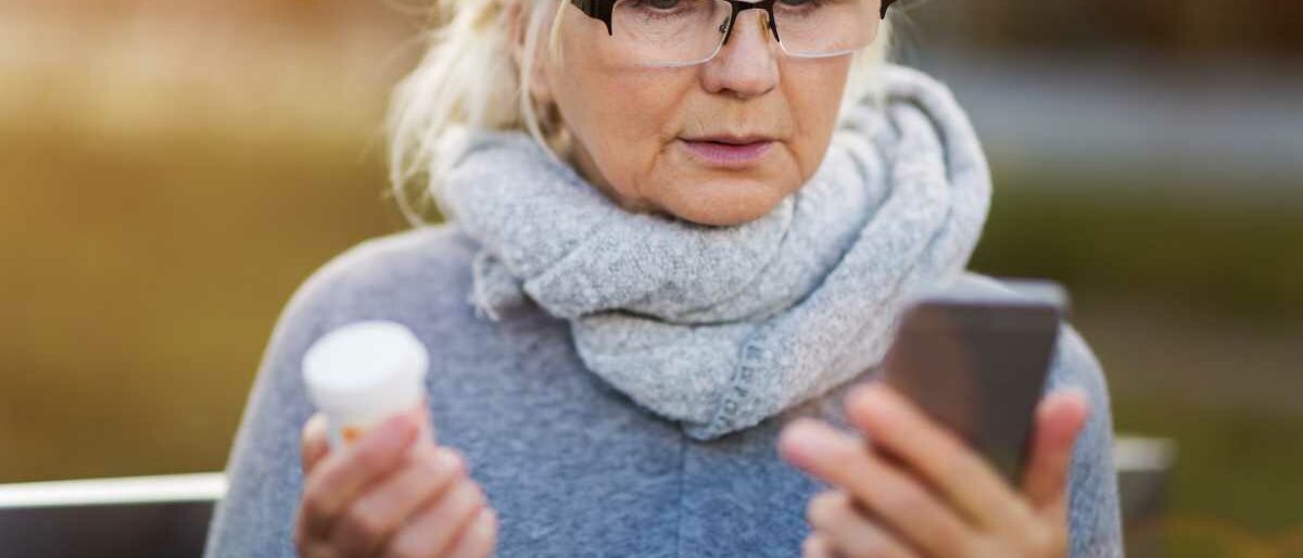 Eine grauhaarige Frau hält in der einen Hand eine Tablettendose, in der anderen ihr Smartphone.
