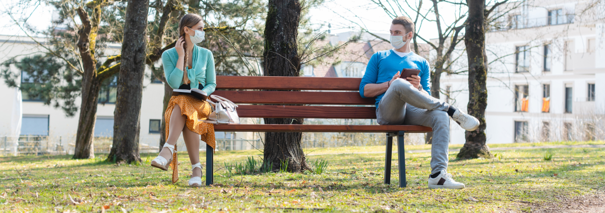 Eine Frau und ein Mann sitzen mit Abstand an den Enden einer Bank, beide tragen Mund-Nasen-Masken.