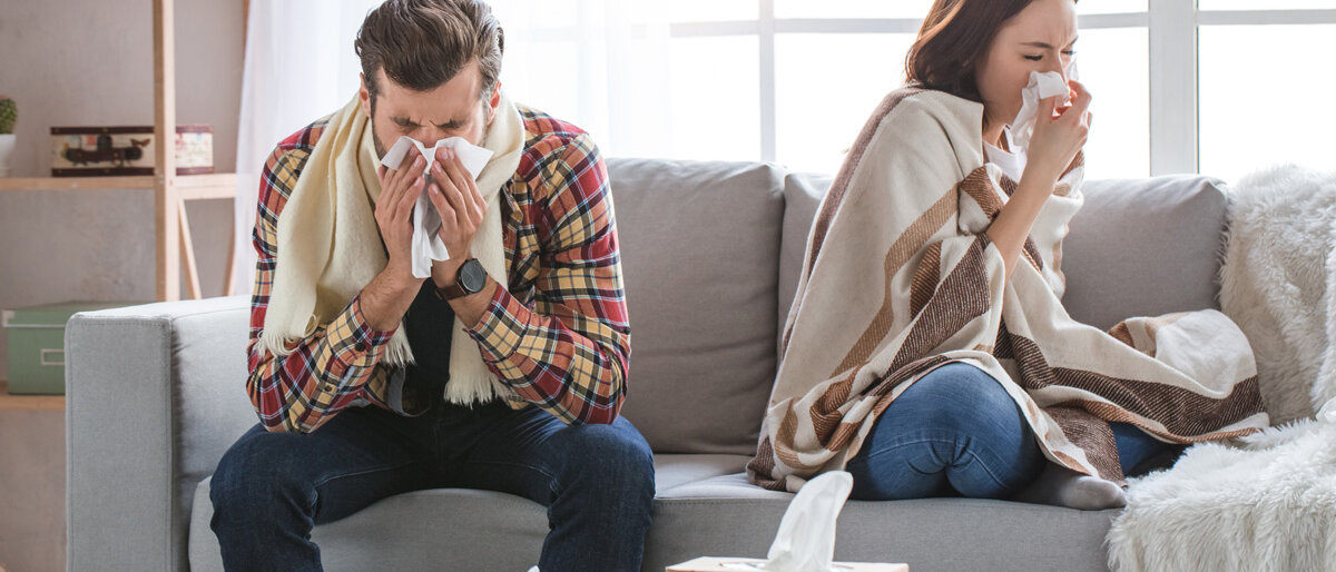 Ein Paar sitzt erkältet auf der Couch, mit Taschentüchern