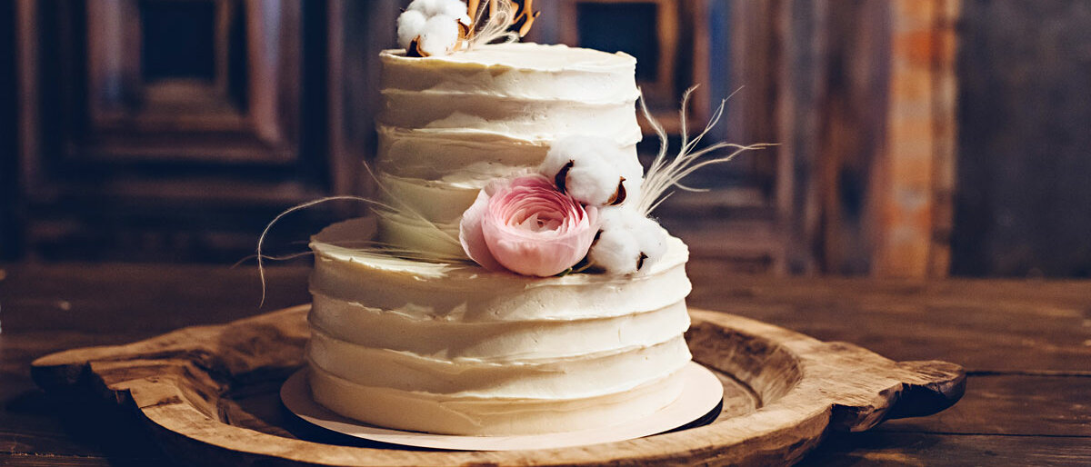 Eine kleine, zweistöckige Hochzeitstorte steht auf einem Holztablett. Sie hat ein Relief aus weißer Buttercreme und ist mit einer Baumwollblüte und einer Pfingstrose geschmückt.